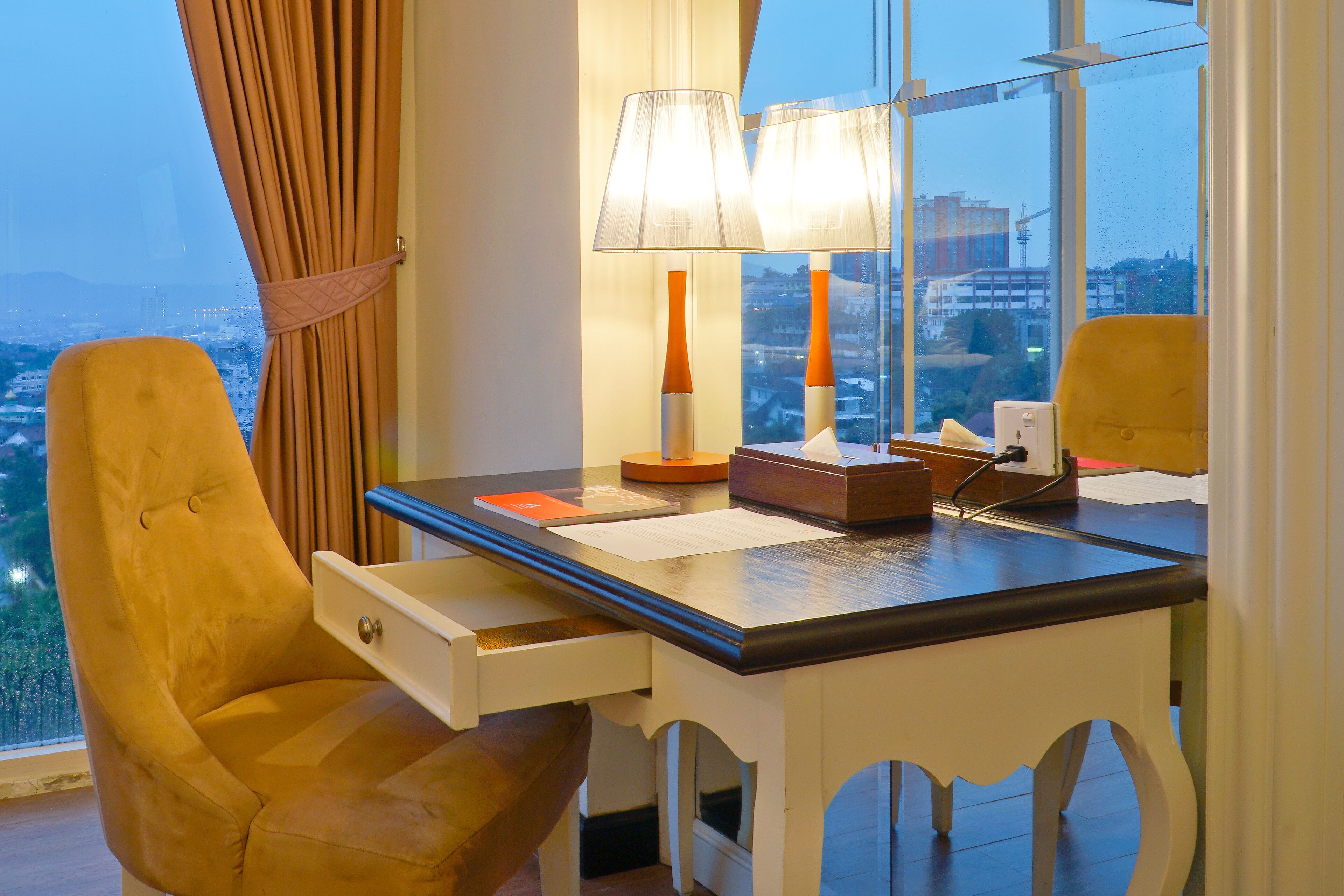 A hotel room with a desk, two lamps, a chair, and a view of the cityscape.