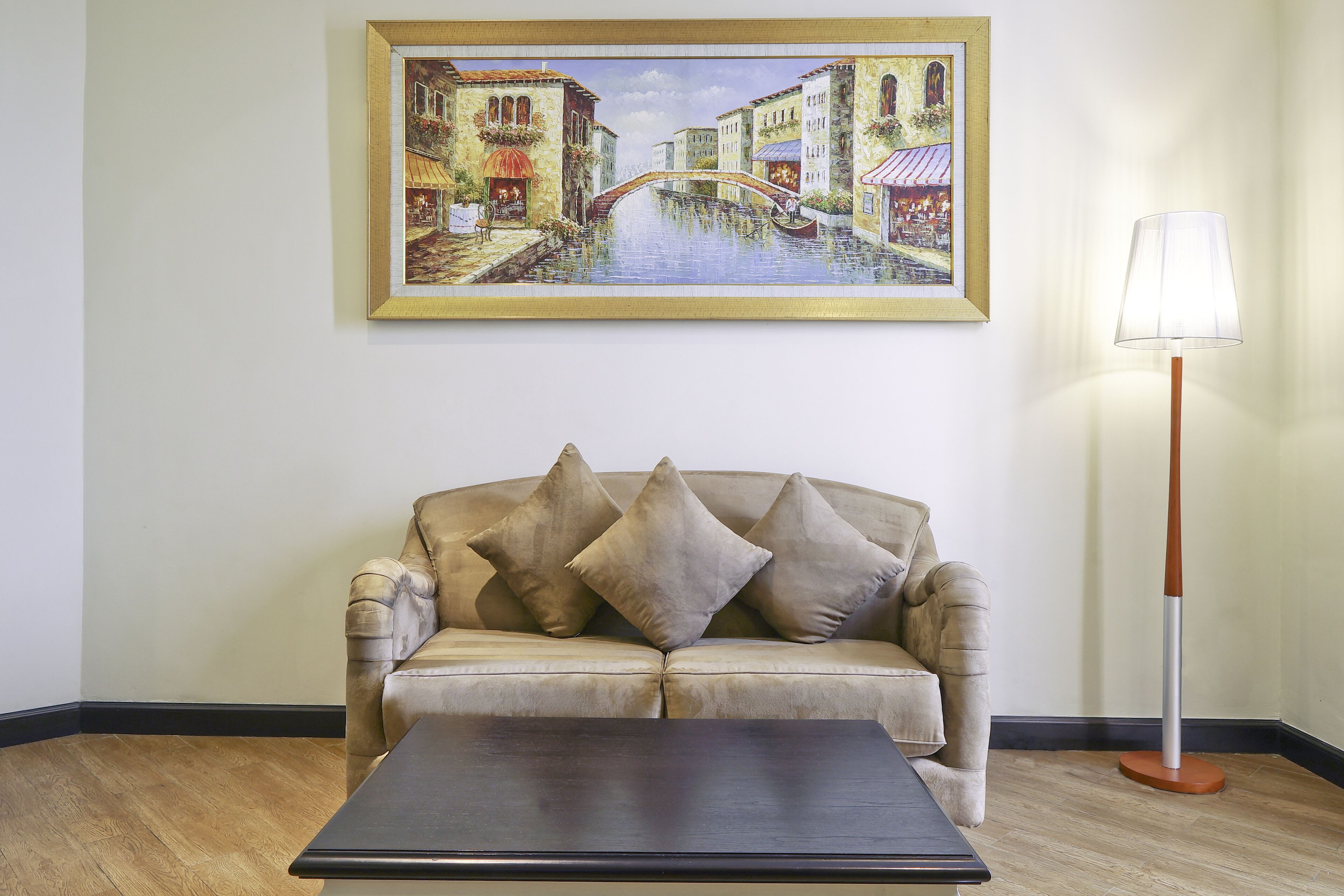 A room with a beige sofa, a framed painting of a canal scene, and a floor lamp.