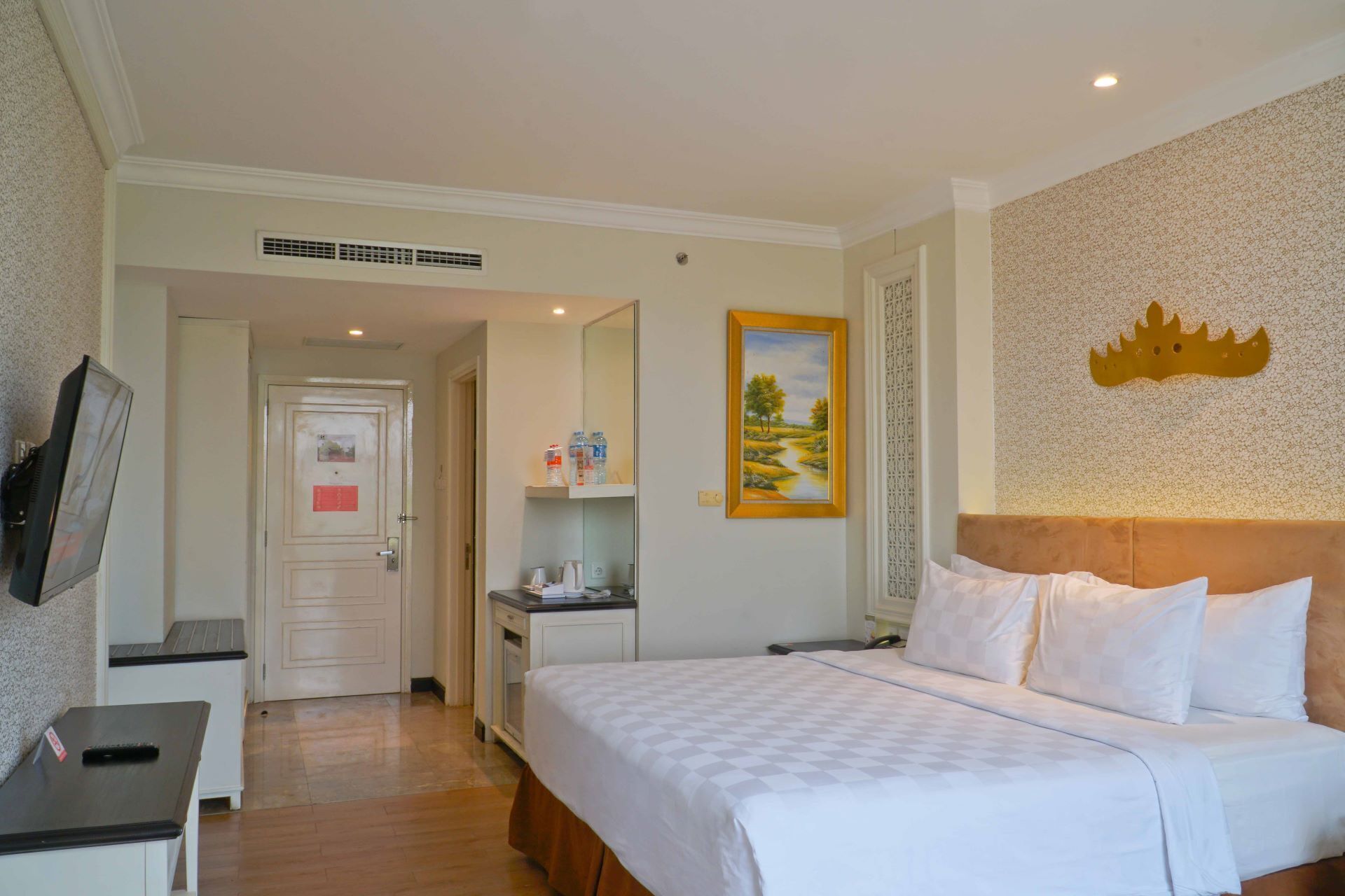 A hotel room with a bed, a desk, a chair, a TV, a painting, and a crown decoration on the wall.