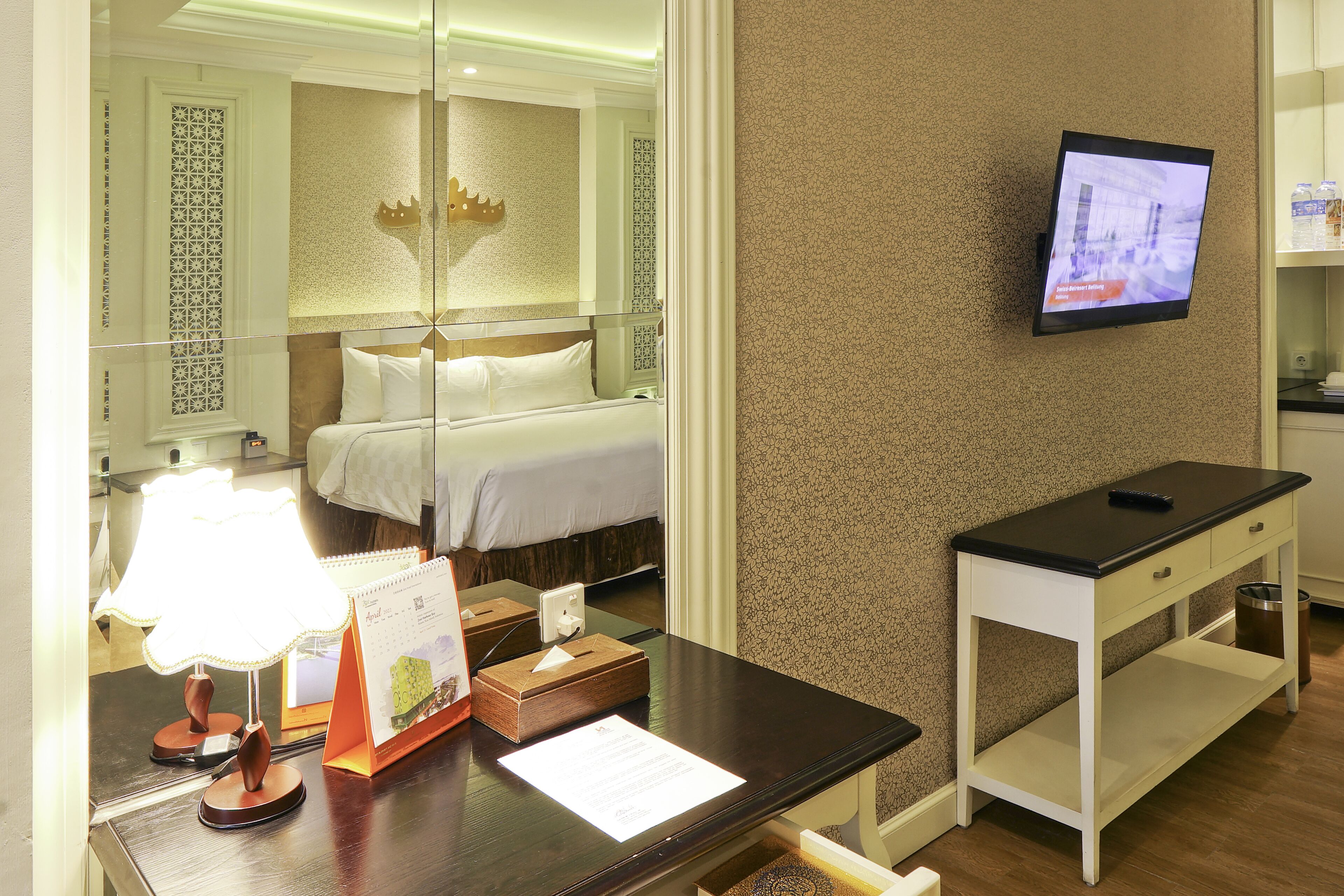Una habitación de hotel con una cama, un escritorio con lámpara, un televisor y una pequeña mesa con un calendario.