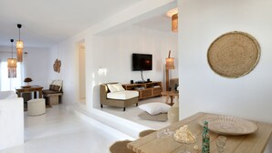 A modern living room with a sofa, coffee table, and TV. There are wicker lamps, a woven wall hanging, and a dining table with glasses and a bottle. - Amalgam Homes Paros (Paros)