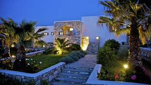 A stone archway leading to a building with a white facade, surrounded by palm trees and a well-lit garden. - Amalgam Homes Paros (Paros)