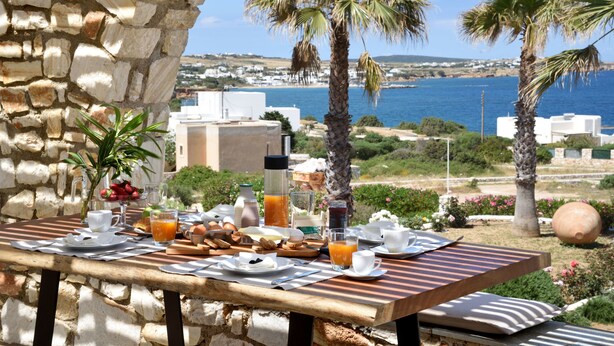 A breakfast table set outdoors with a stone wall backdrop, palm trees, and a view of the sea and buildings. - Amalgam Homes Paros (Paros)