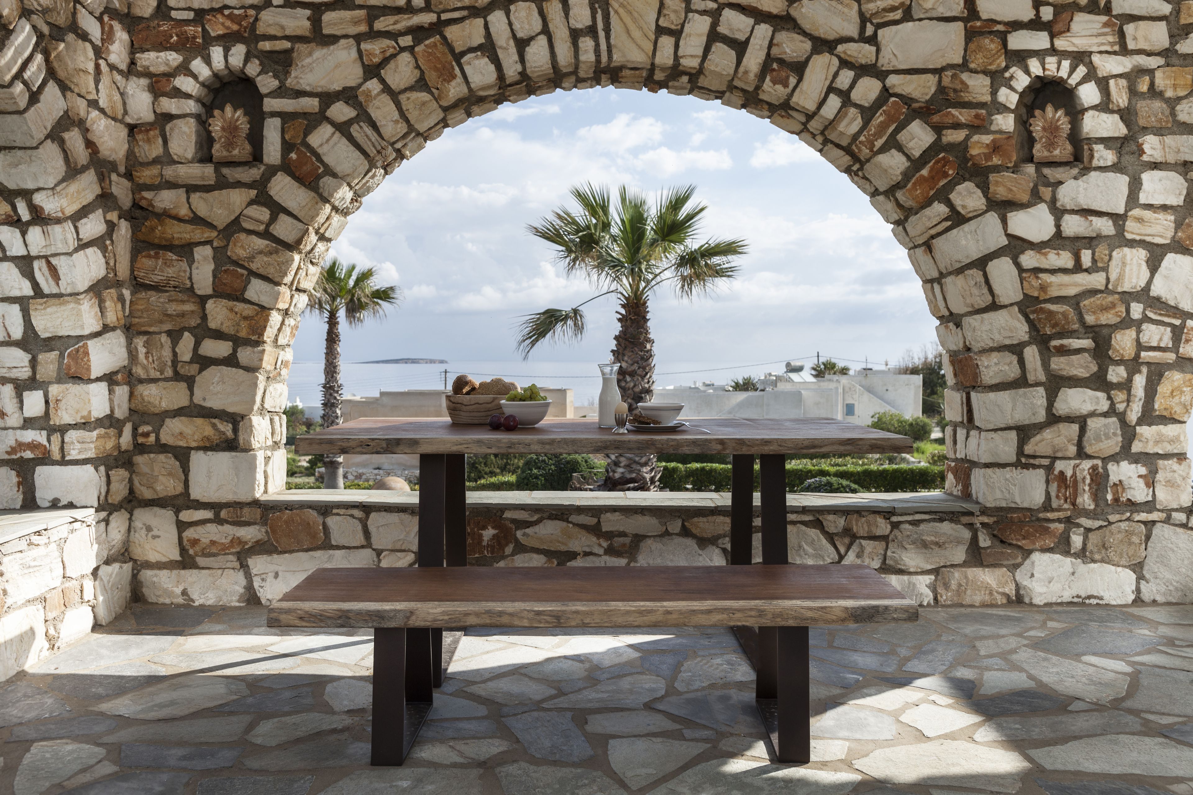 Un arco de piedra con una mesa y un banco, con vistas a una palmera y edificios.
