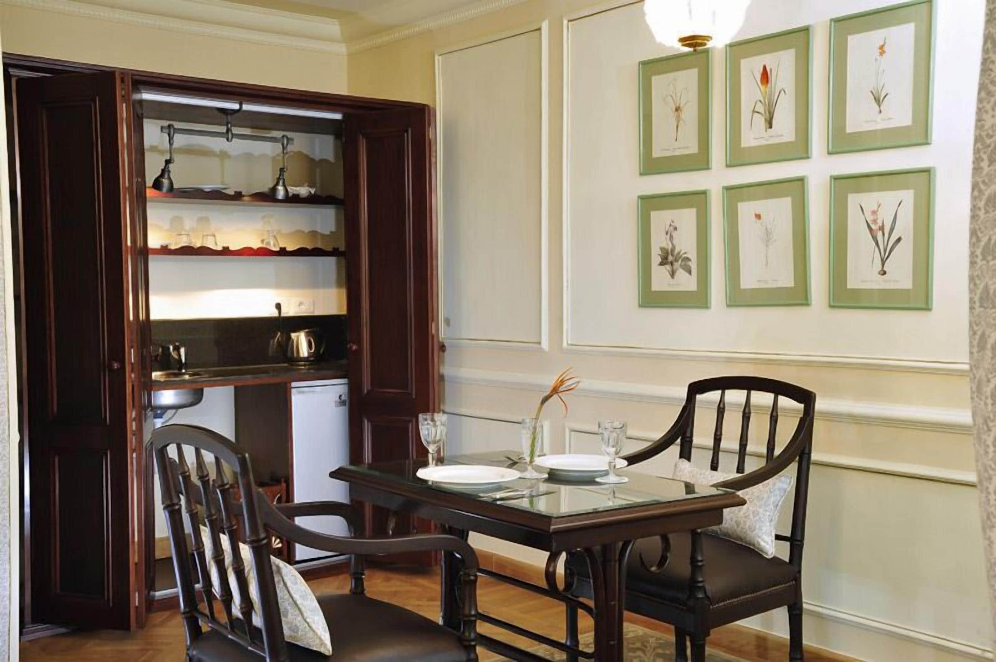 A dining area with a glass-top table, two chairs, a small cabinet with shelves, and framed botanical illustrations on the wall.