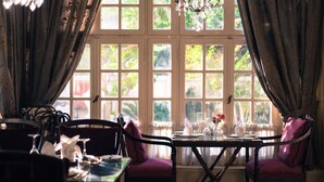 A dining area with a table set for two, surrounded by chairs, and a view of greenery outside through large windows. - Villa Belle Epoque (Cairo)