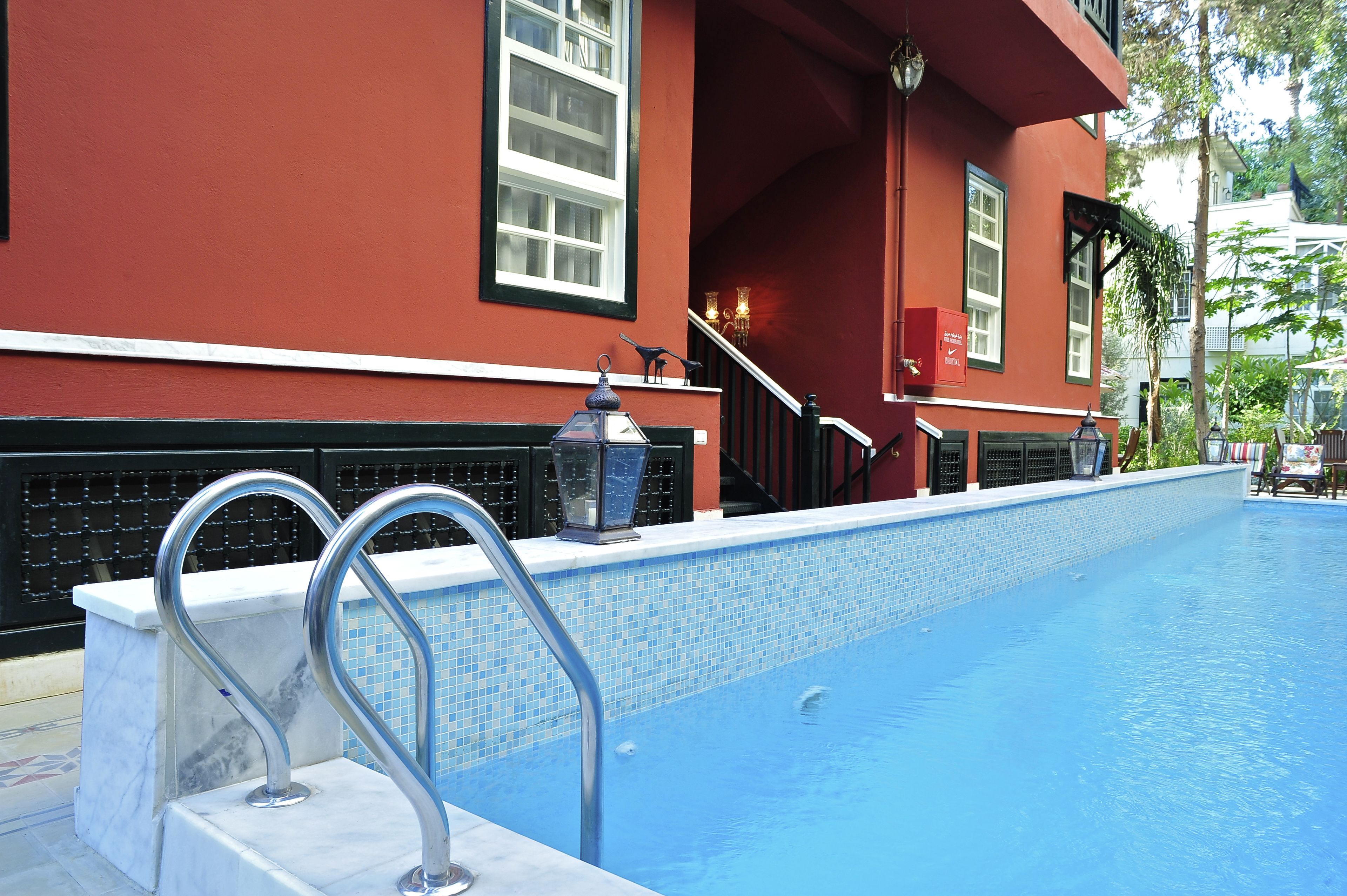 Une piscine dont le bord est recouvert de carreaux, avec une rambarde métallique, et un bâtiment rouge orné de moulures blanches, avec un escalier menant à un balcon.