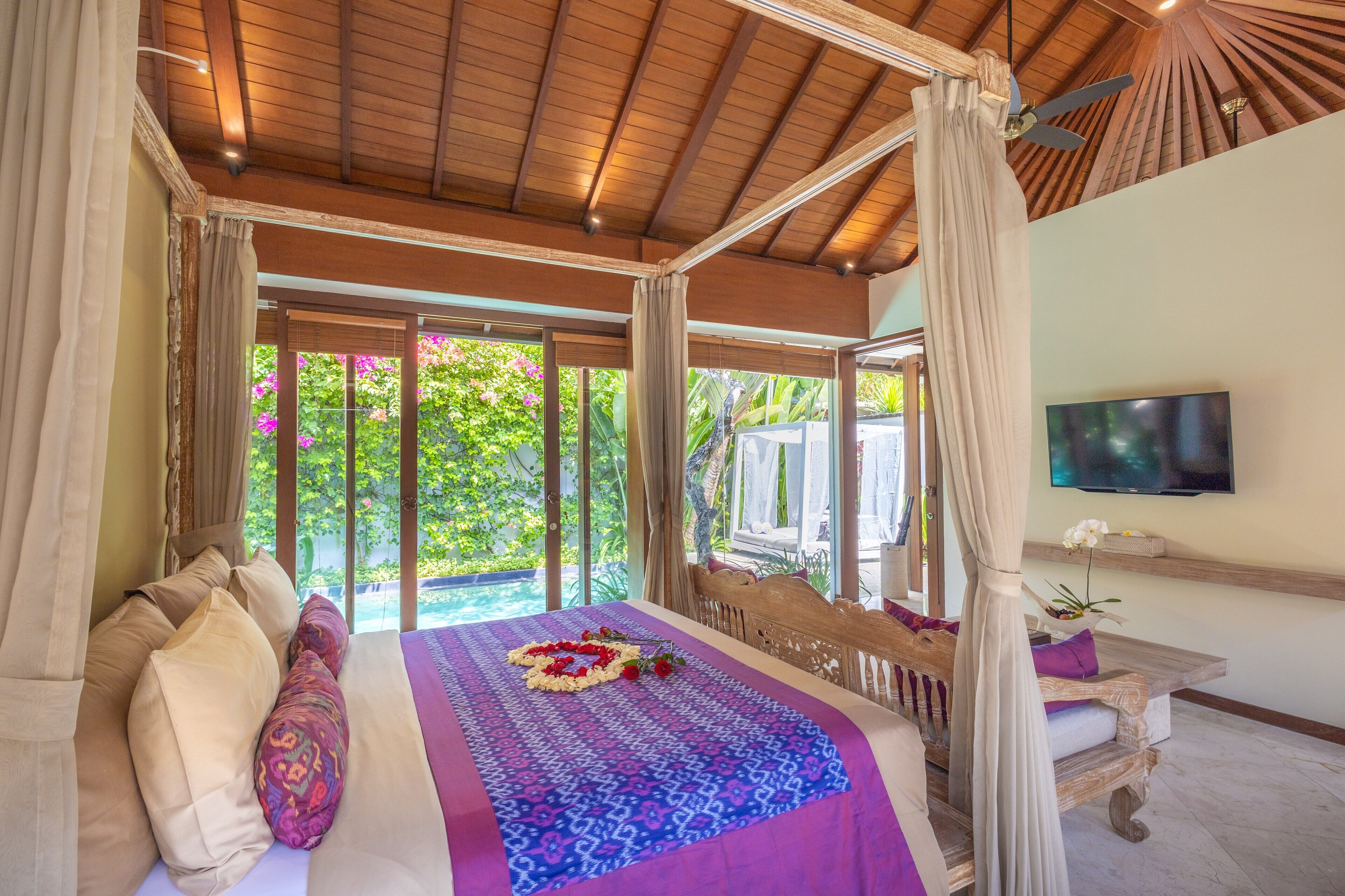 A bedroom with a canopy bed, a television on the wall, and a view of greenery outside.