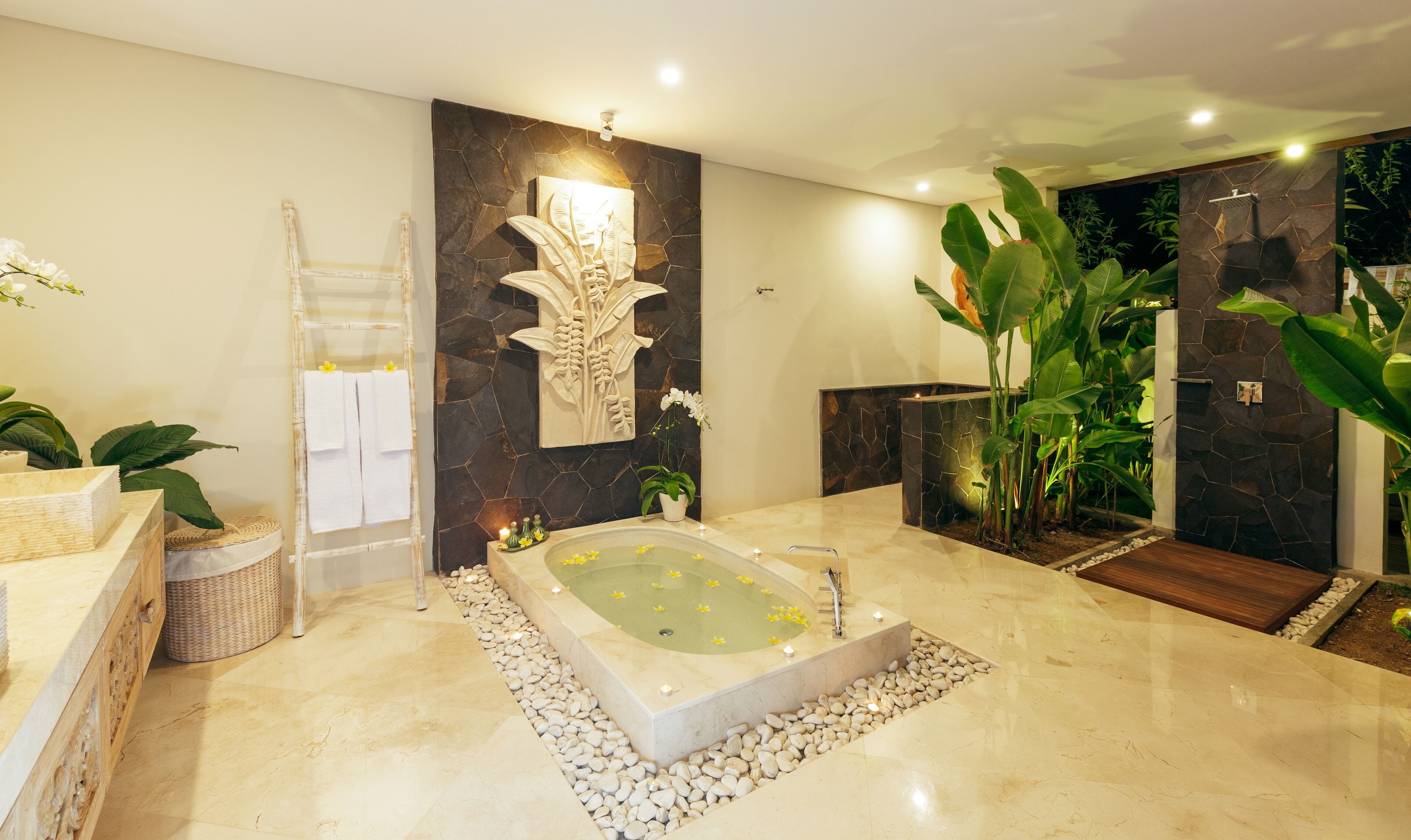 A modern bathroom with a freestanding bathtub, a stone wall with a leaf design, and various plants.
