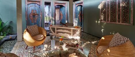 Une pièce spacieuse avec une table basse en verre, deux fauteuils en osier, un canapé à motifs et un panneau décoratif mural.