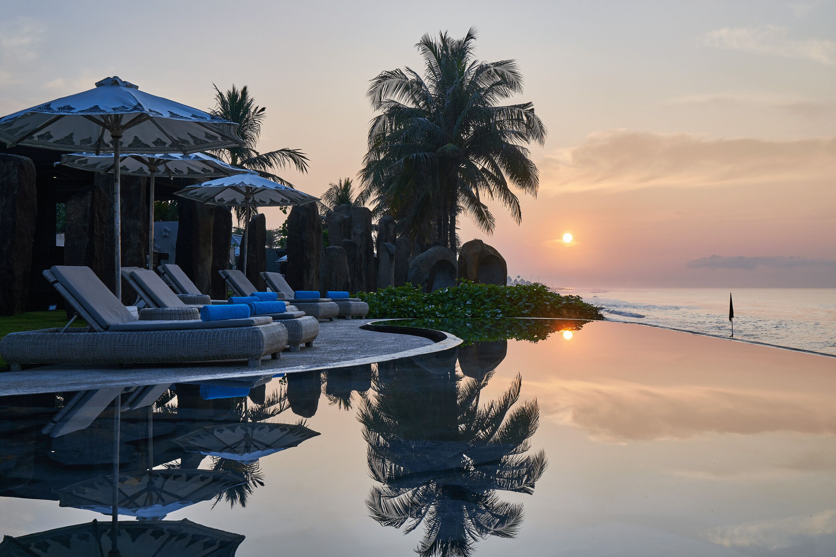 Una piscina infinita con sillas de descanso y sombrillas, palmeras y vistas al atardecer.