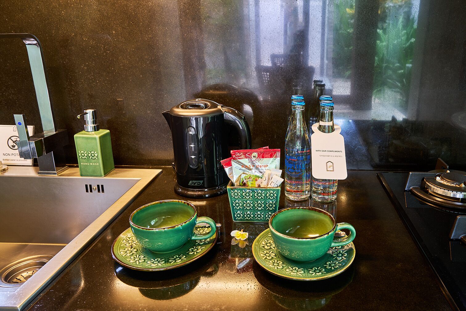 A kitchen counter with a coffee maker, two cups, a bottle of water, a hand wash soap dispenser, and a kettle.
