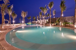 A large, curved swimming pool at night, surrounded by palm trees and lounge chairs.