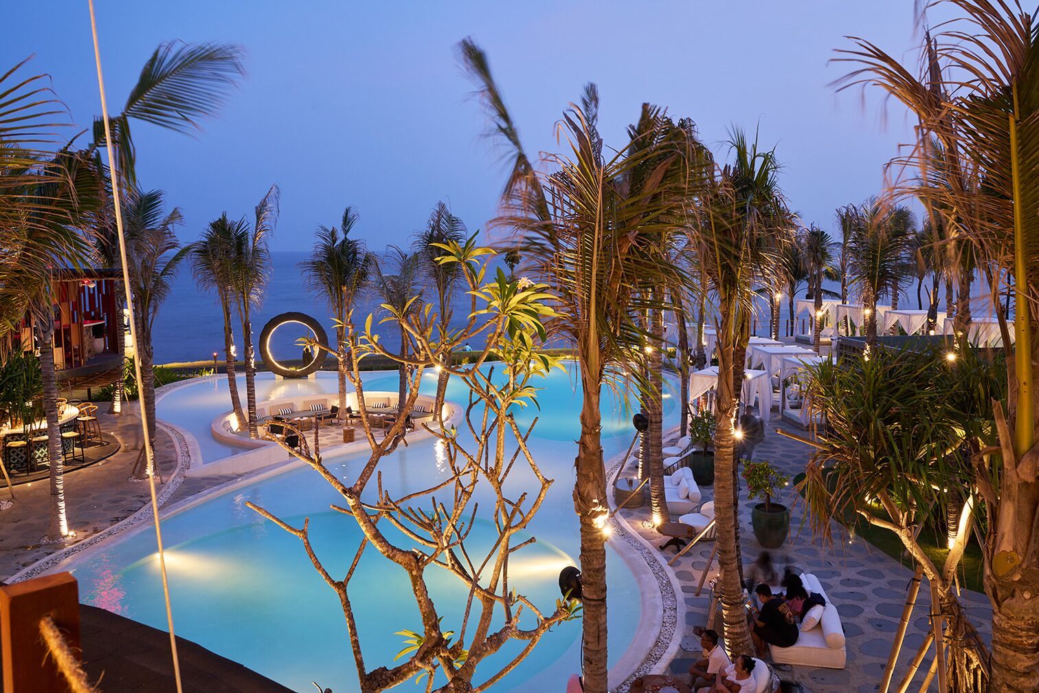 A pool area with lounge chairs and palm trees, surrounded by a pool deck and a view of the ocean.