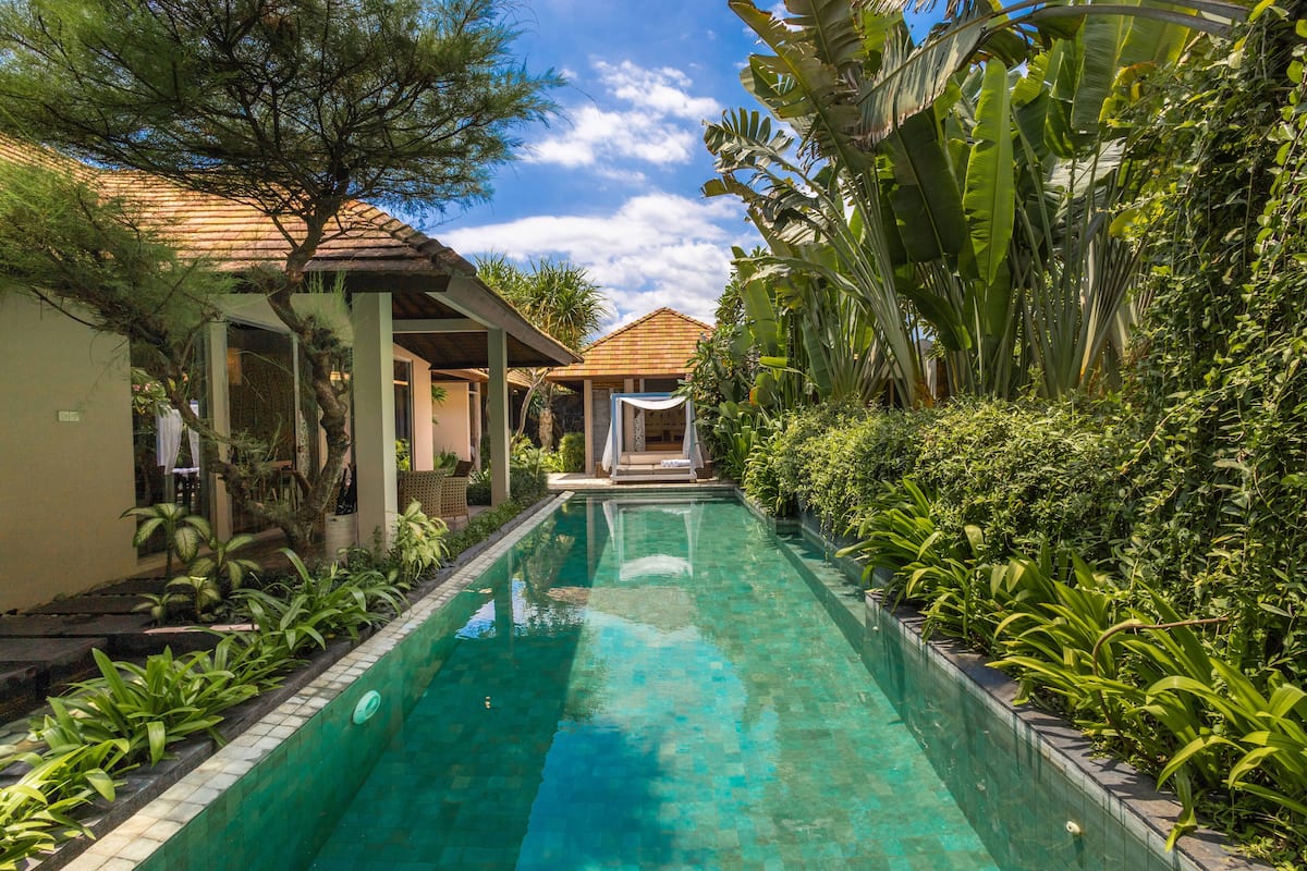 A swimming pool with a tiled edge, surrounded by lush greenery and a building with a tiled roof.