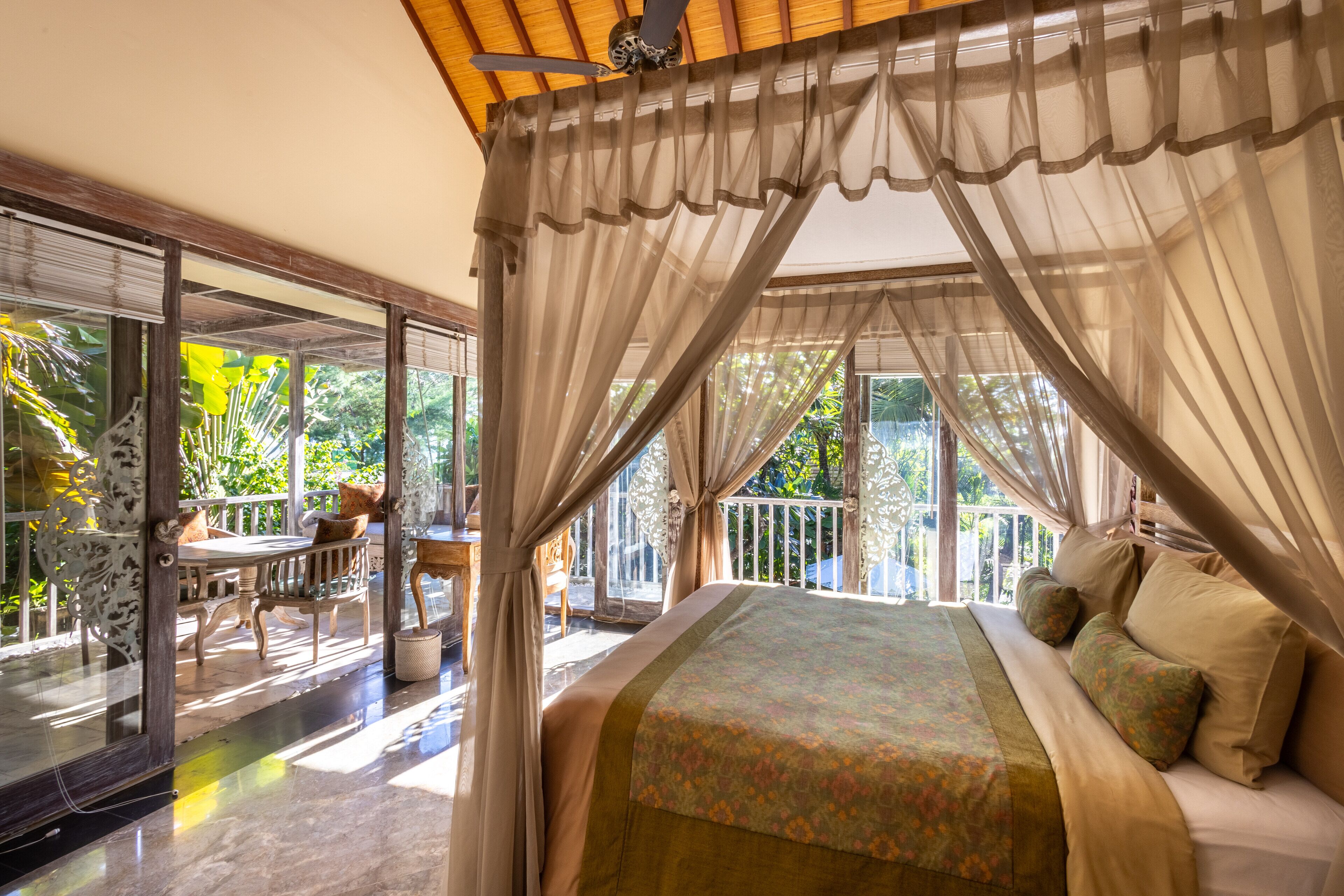 A bedroom with a canopy bed, a balcony with a table and chairs, and a view of greenery outside.