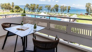 A balcony with a table and chairs, overlooking a pool area with umbrellas and a view of the sea and mountains. - Kerkyra Blue Hotel & Spa by Louis Hotels (Corfu)