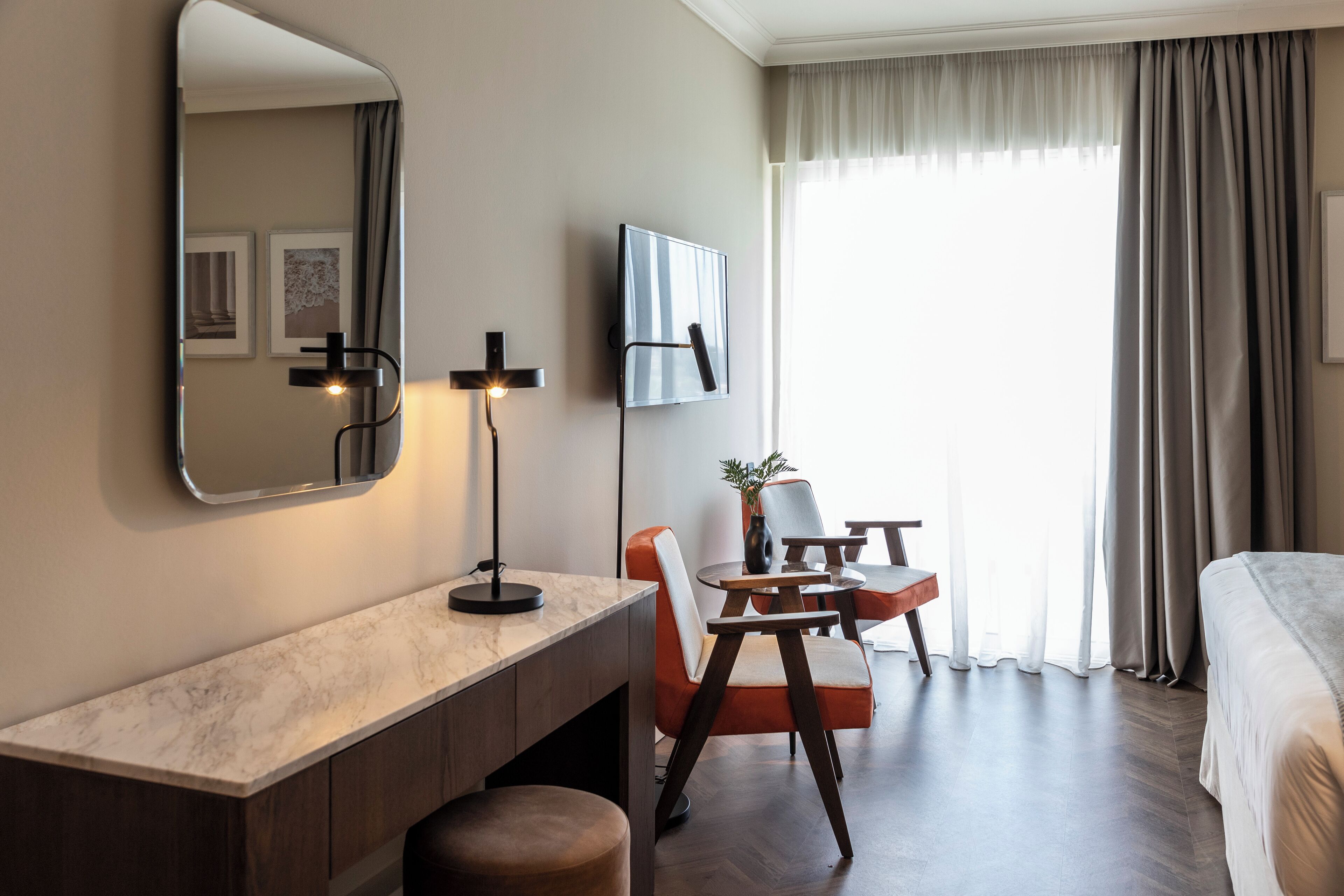 Habitación de hotel con mesita de noche de mármol, un espejo grande, televisor de pantalla plana en la pared, un escritorio de madera con una silla y una cama con cabecera.