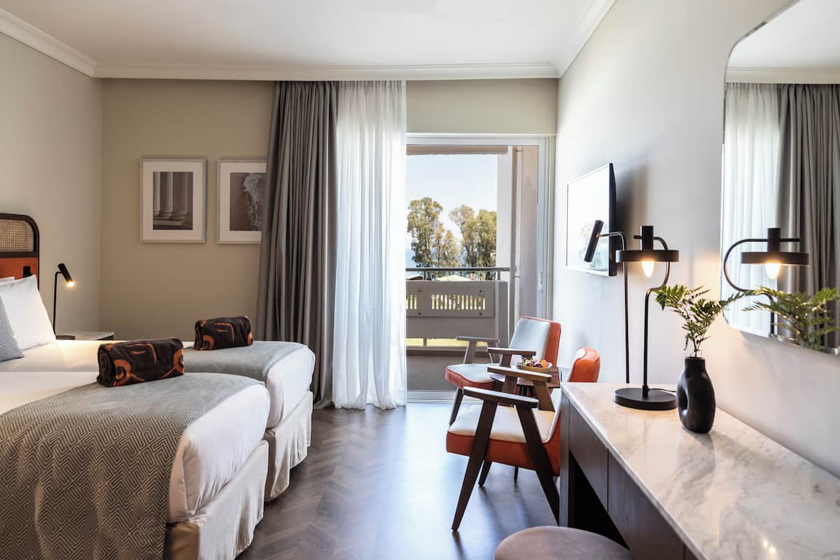 A hotel room with two beds, a desk, a chair, a TV, and a balcony with a view.