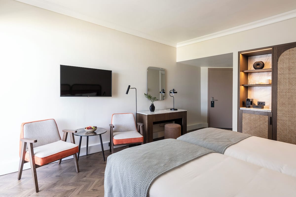 A hotel room with a bed, two chairs, a desk with a mirror, a small table with a bowl, and a shelf with decorative items.