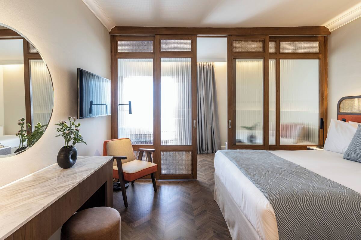 A hotel room with a bed, a desk with a chair, a TV, and a large window with curtains.