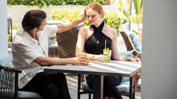 A man and a woman sitting at an outdoor table with drinks, surrounded by wicker furniture and greenery.