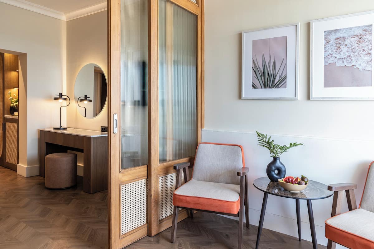 A modern hotel room with a wooden door, a round mirror, two chairs, a small table with a vase and fruit bowl, and framed artwork on the wall.