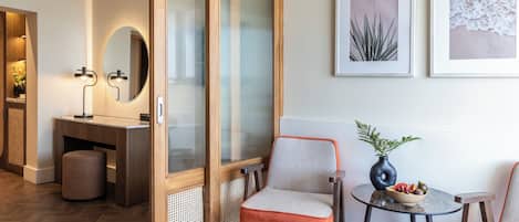 Habitación de hotel moderna con puerta de madera, espejo redondo, dos sillas, una mesita con un florero y frutero, y obras de arte enmarcadas en la pared.