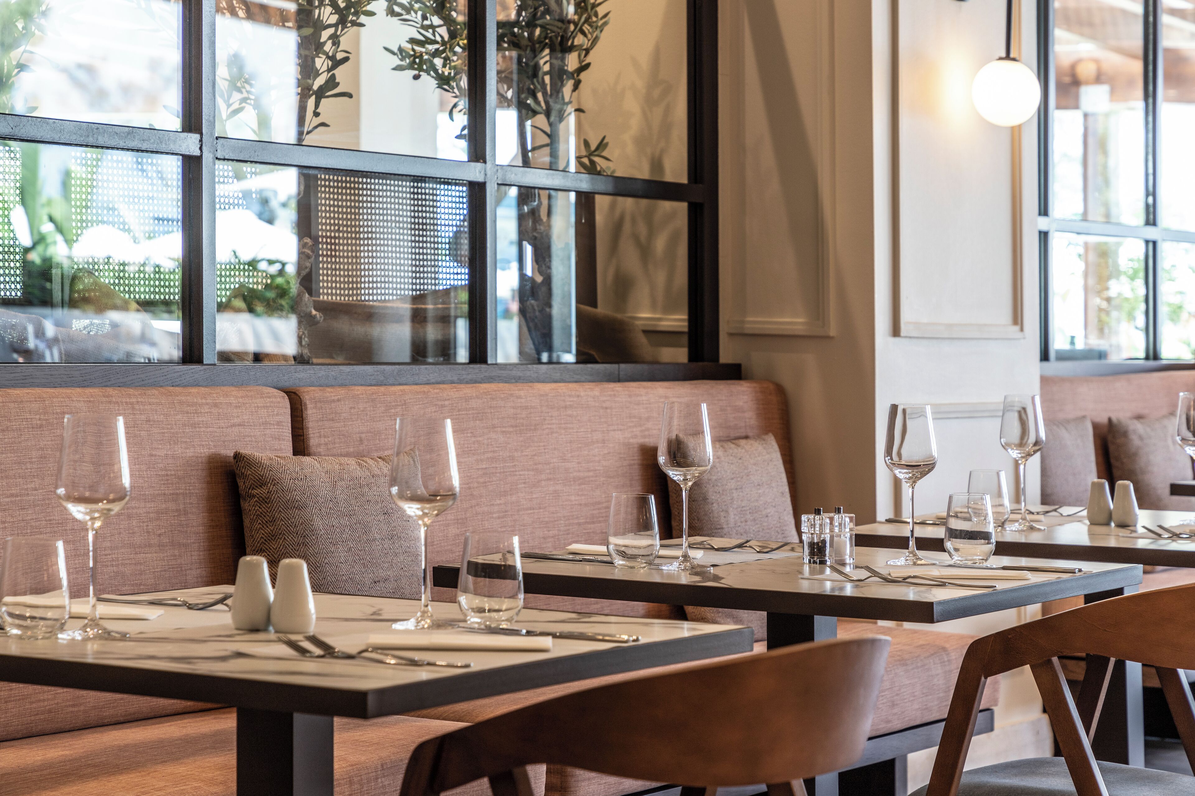 A restaurant with tables set for dining, a seating area with cushions, and a view through a window.