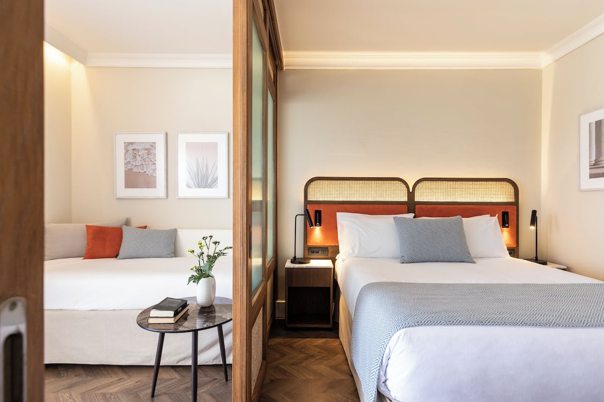 A hotel room with two beds, a small table with a book and a plant, and framed artwork on the walls.