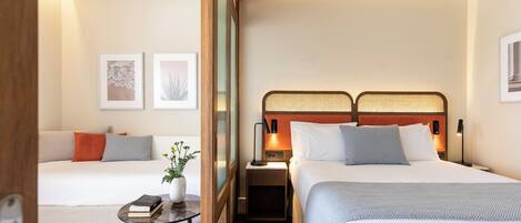 A hotel room with two beds, a small table with a book and a plant, and framed artwork on the walls.