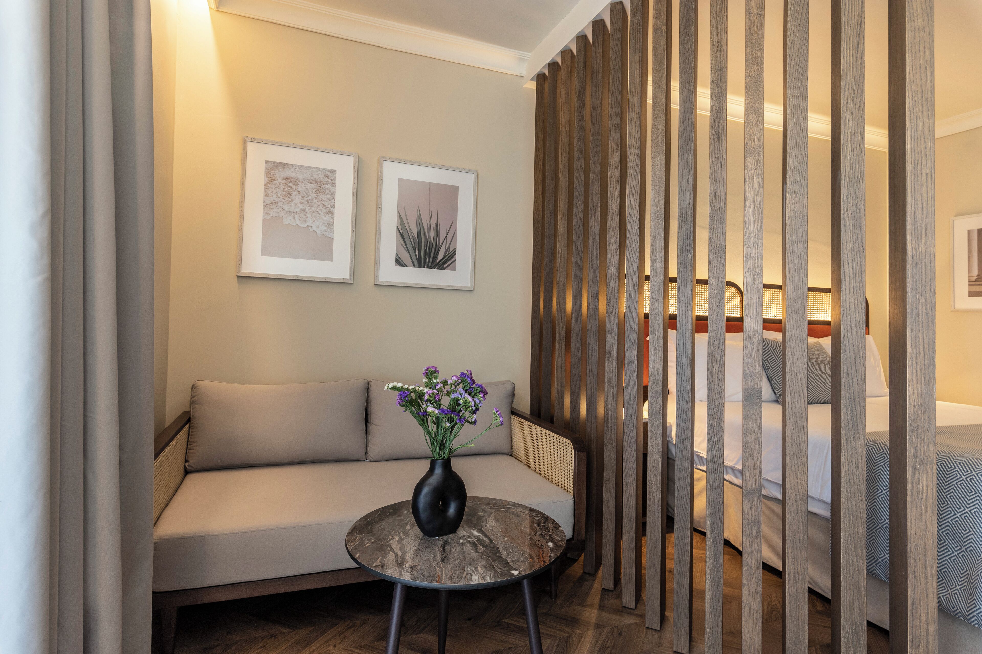 Ein modernes Hotelzimmer mit einer Couch, einem kleinen runden Tisch mit Blumenvase und einem Bett mit gemustertem Überwurf.
