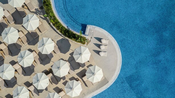 An aerial view of a resort with multiple sun loungers and umbrellas arranged along a curved poolside. - Kerkyra Blue Hotel & Spa by Louis Hotels (Corfu)