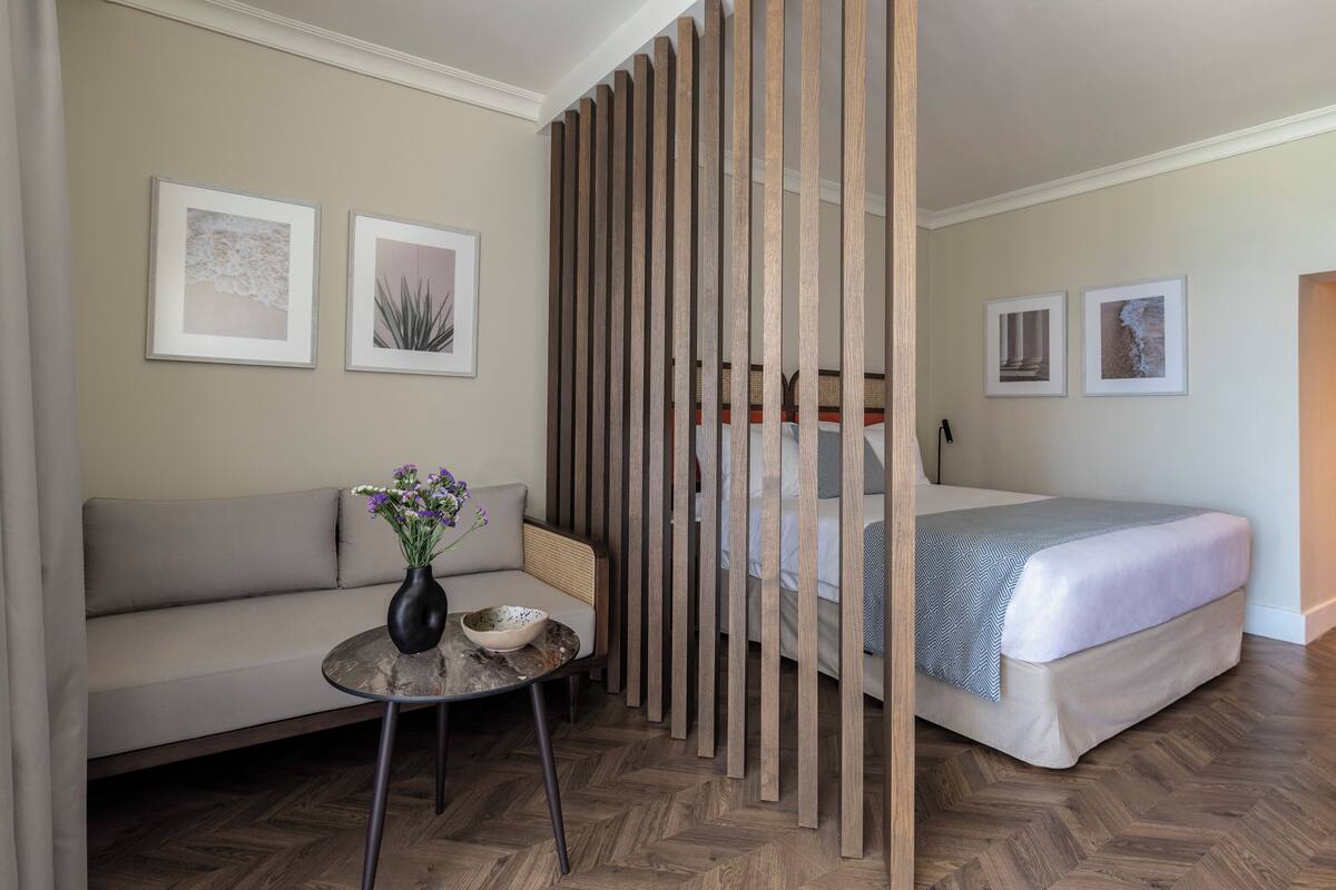 A hotel room with a bed, a sofa, a small table with a vase, and two framed pictures on the wall.