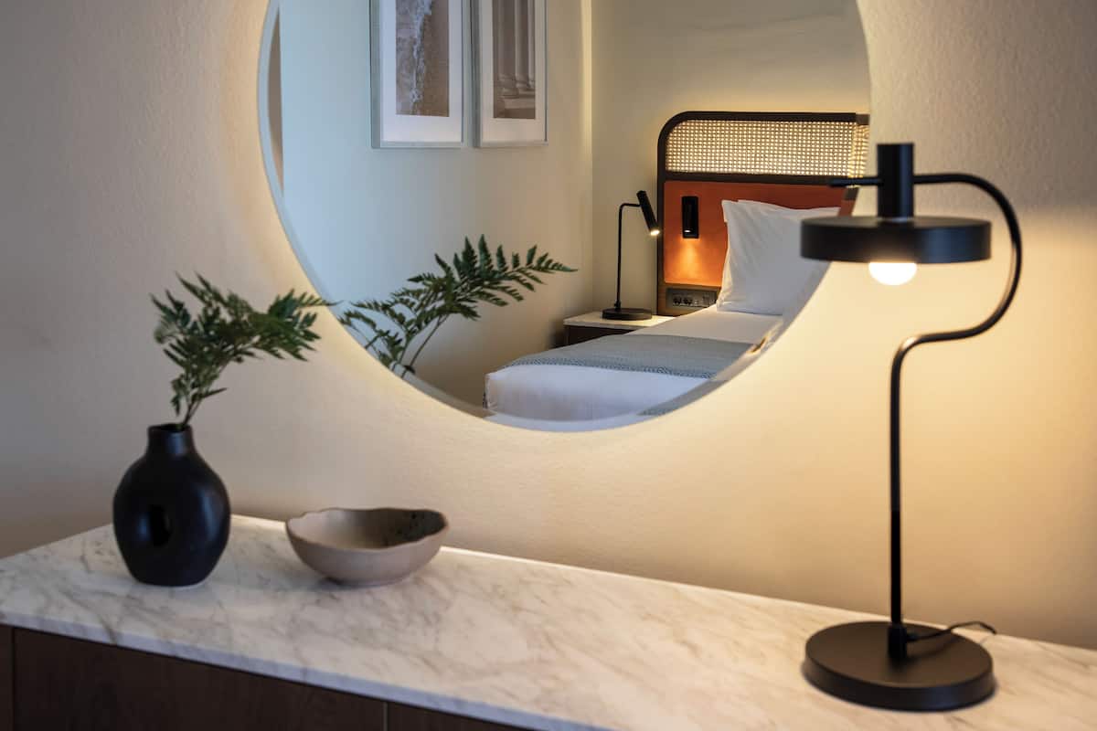 A modern bedroom with a circular mirror, a bedside lamp, a vase, and a bowl on a marble countertop.