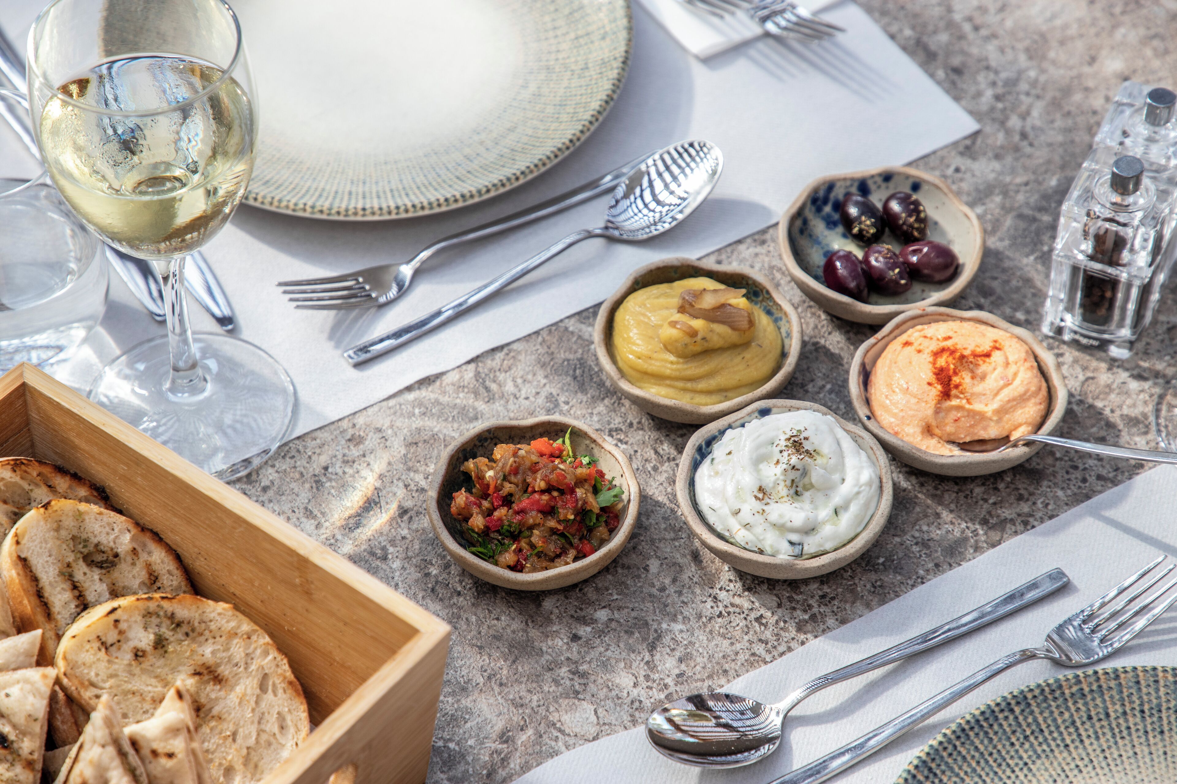 Een gedekte tafel met diverse soorten eten, waaronder bakjes met dips en olijven, een glas witte wijn en een houten broodkist.
