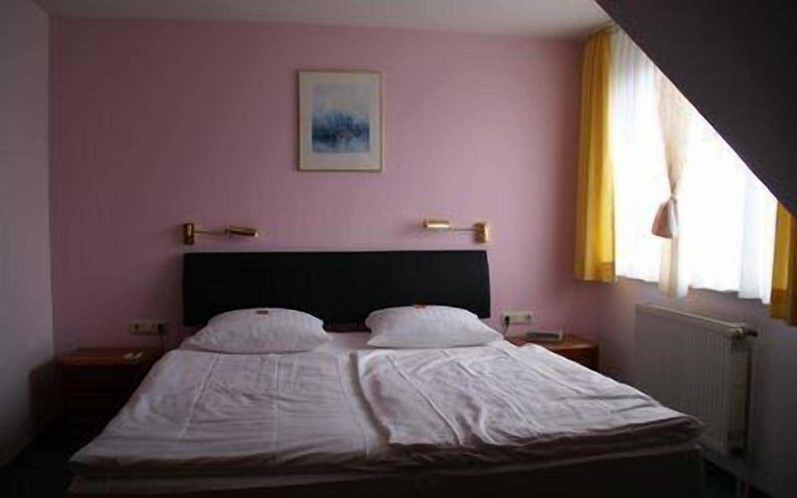 Ein Einzelbett in einem Zimmer mit rosafarbenen Wänden, einem gerahmten Bild an der Wand und einem Fenster mit Vorhängen.