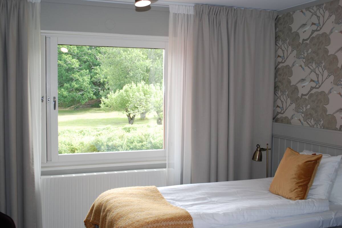 Une chambre avec une grande fenêtre, un lit recouvert de draps blancs et orné d’un coussin marron, et un mur décoré d’un motif floral.