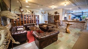 Un vaste salon doté de canapés en cuir, d’une table basse et d’une cheminée. On peut également apercevoir, au fond, des animaux empaillés et un coin bar.