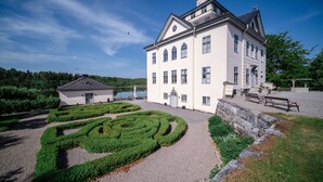 Un grand bâtiment blanc à la façade symétrique, un jardin à la française avec des haies géométriques et un mur en pierre.