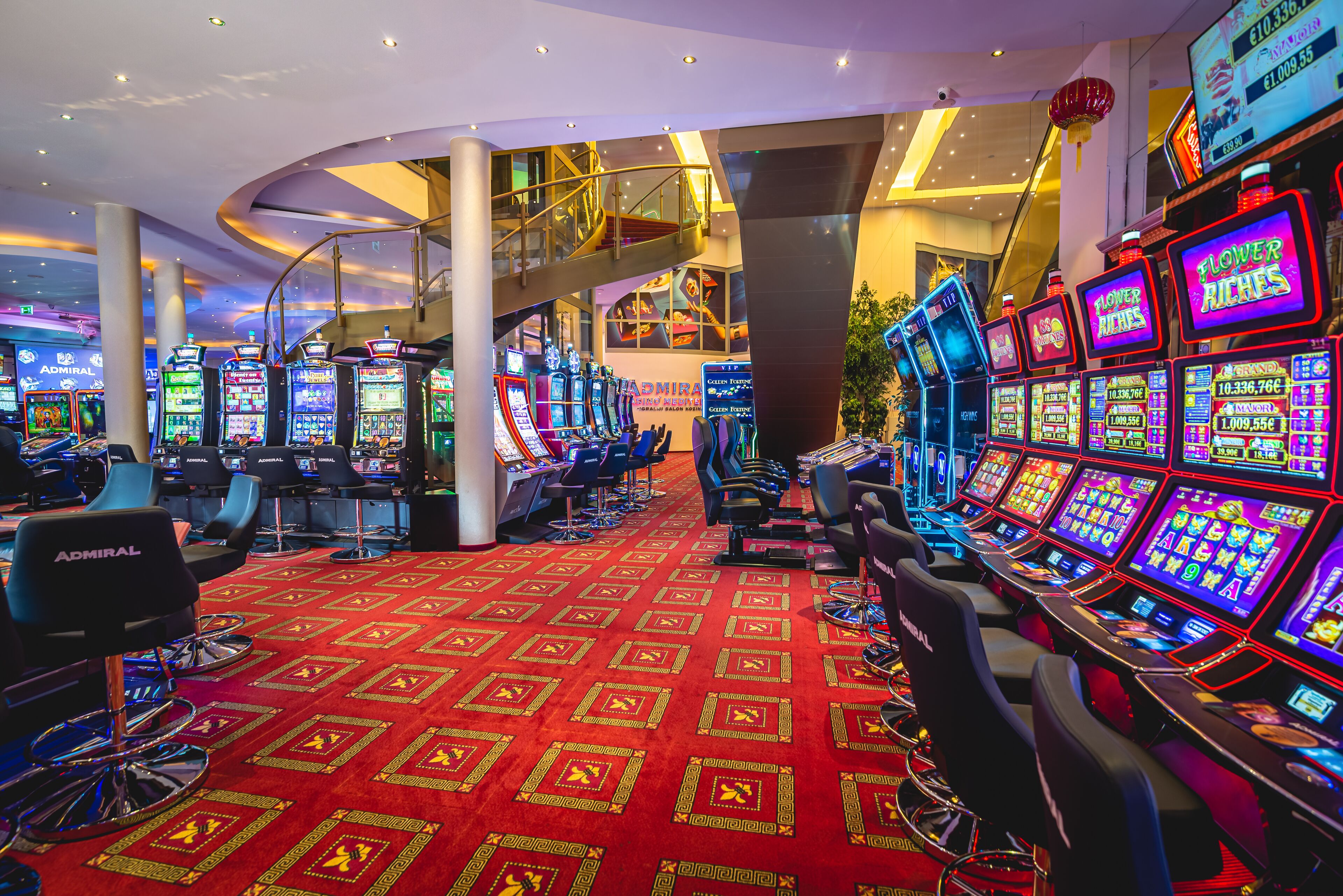 Un casinò con diverse slot machine, un pavimento ricoperto di tappeto rosso e una scala che conduce a un livello superiore.