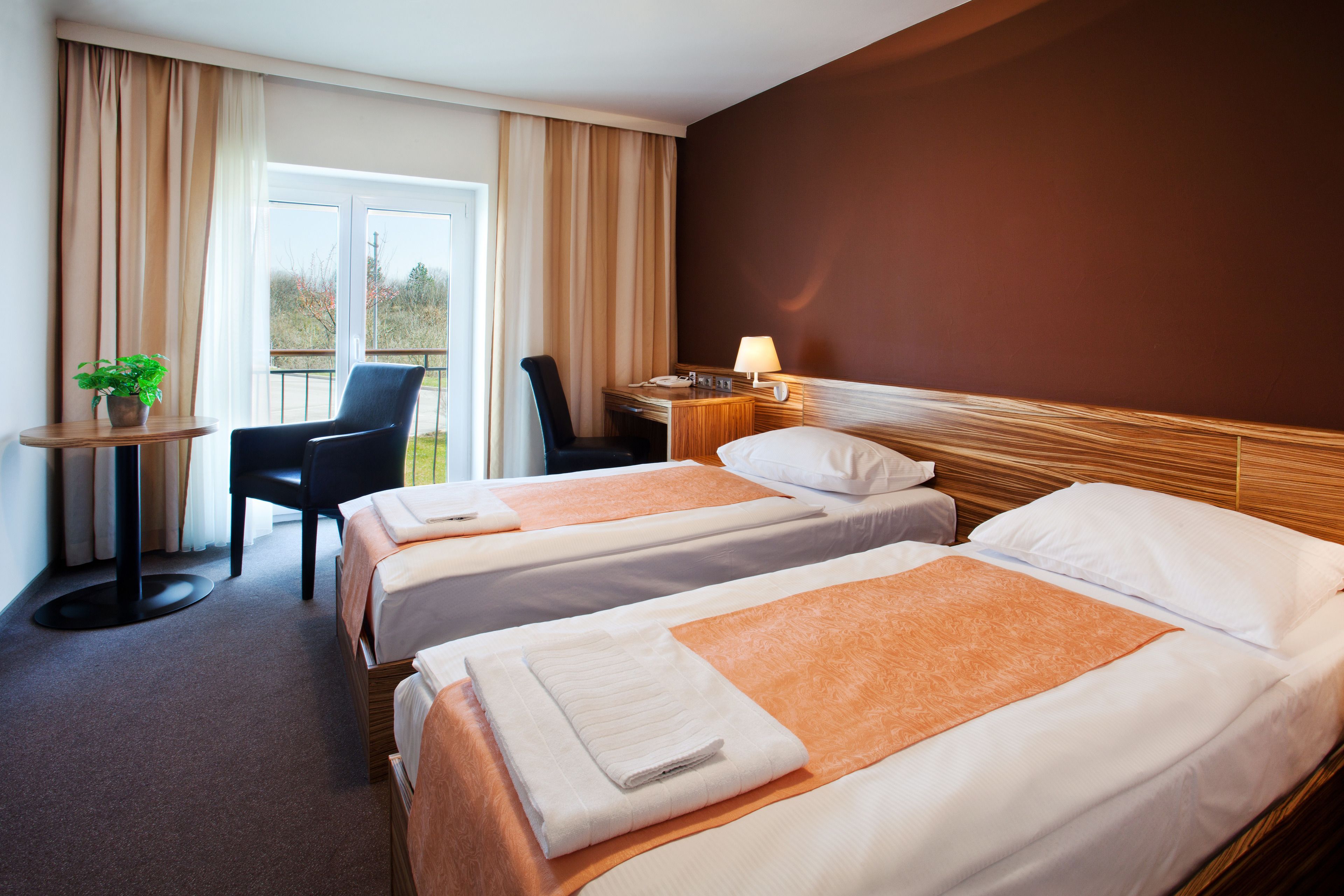 A hotel room with two beds, a desk, a chair, a table with a plant, and a window with curtains.