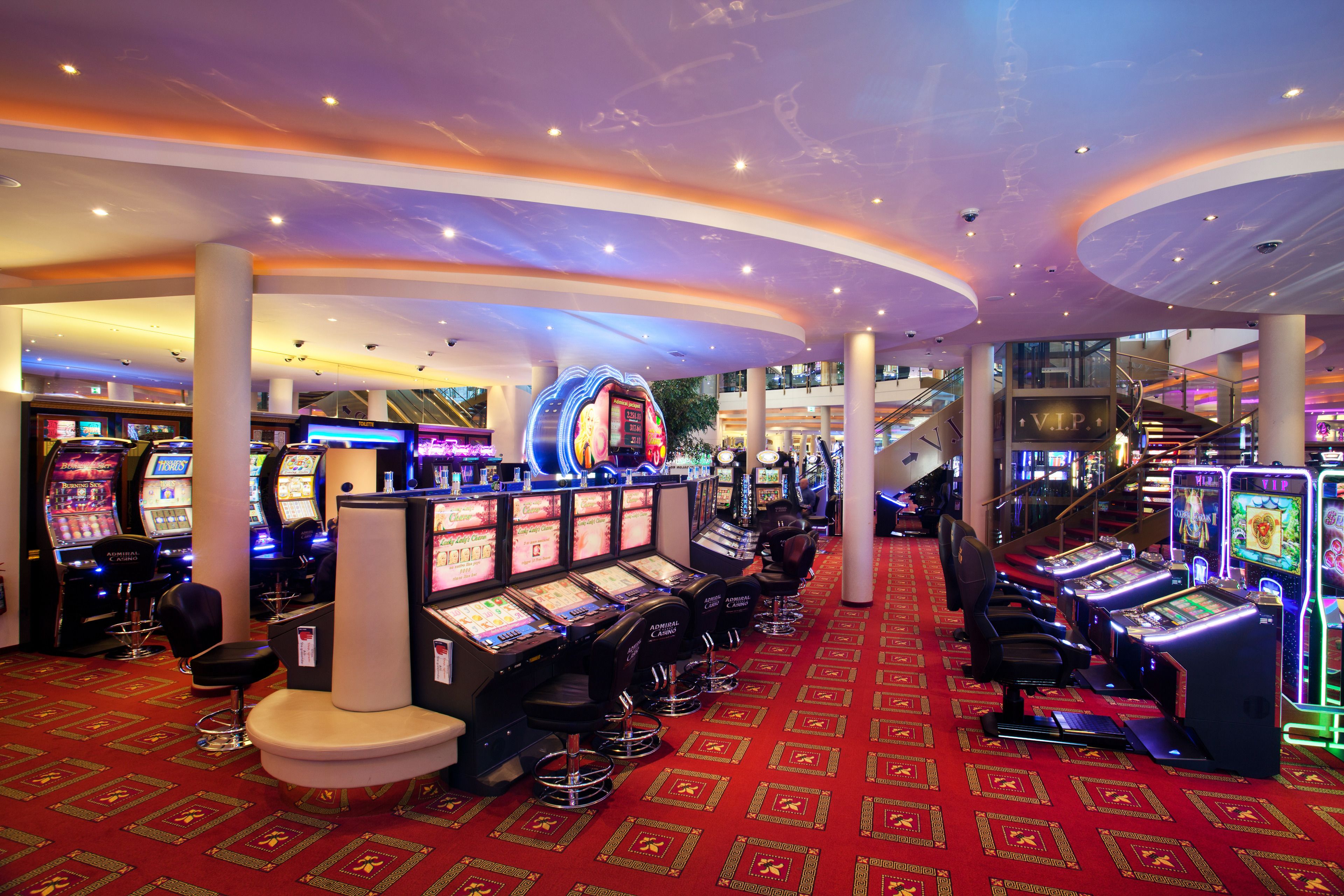 Piso de casino con múltiples máquinas tragamonedas, taburetes de bar y una zona alfombrada de color rojo.