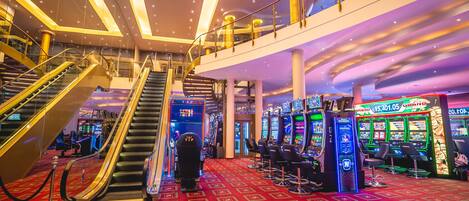 Interior de un casino con alfombra roja, máquinas tragamonedas y una escalera mecánica.