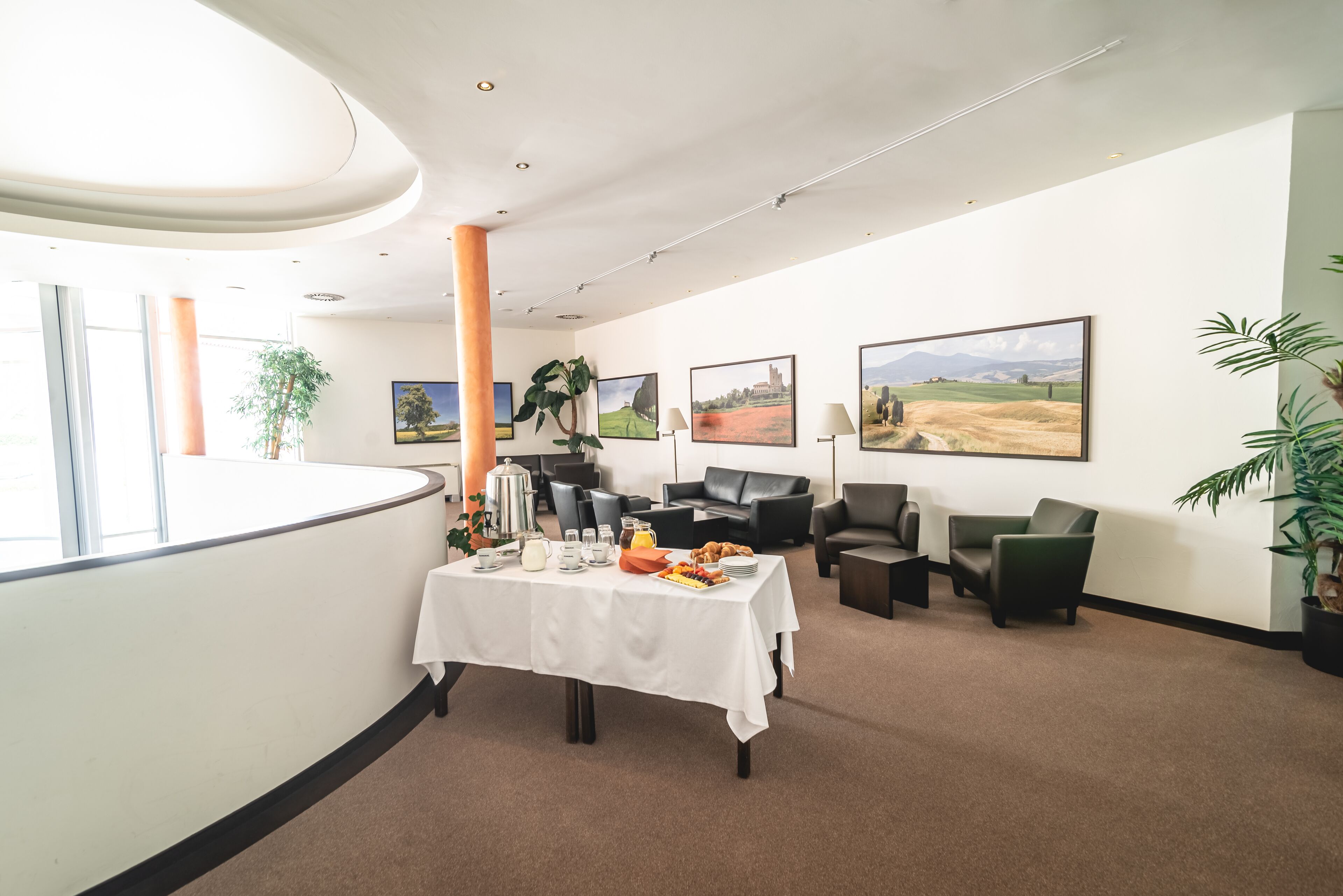 A modern hotel lobby with a reception desk, a buffet table with breakfast items, comfortable seating, and large landscape paintings on the walls.