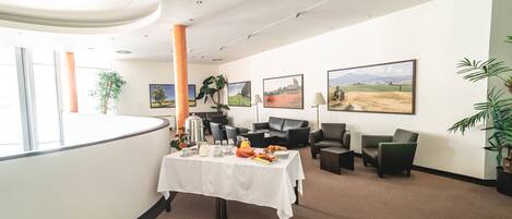 Un moderno vestíbulo de hotel con mostrador de recepción, una mesa de buffet con alimentos para el desayuno, asientos cómodos y grandes cuadros con paisajes en las paredes.