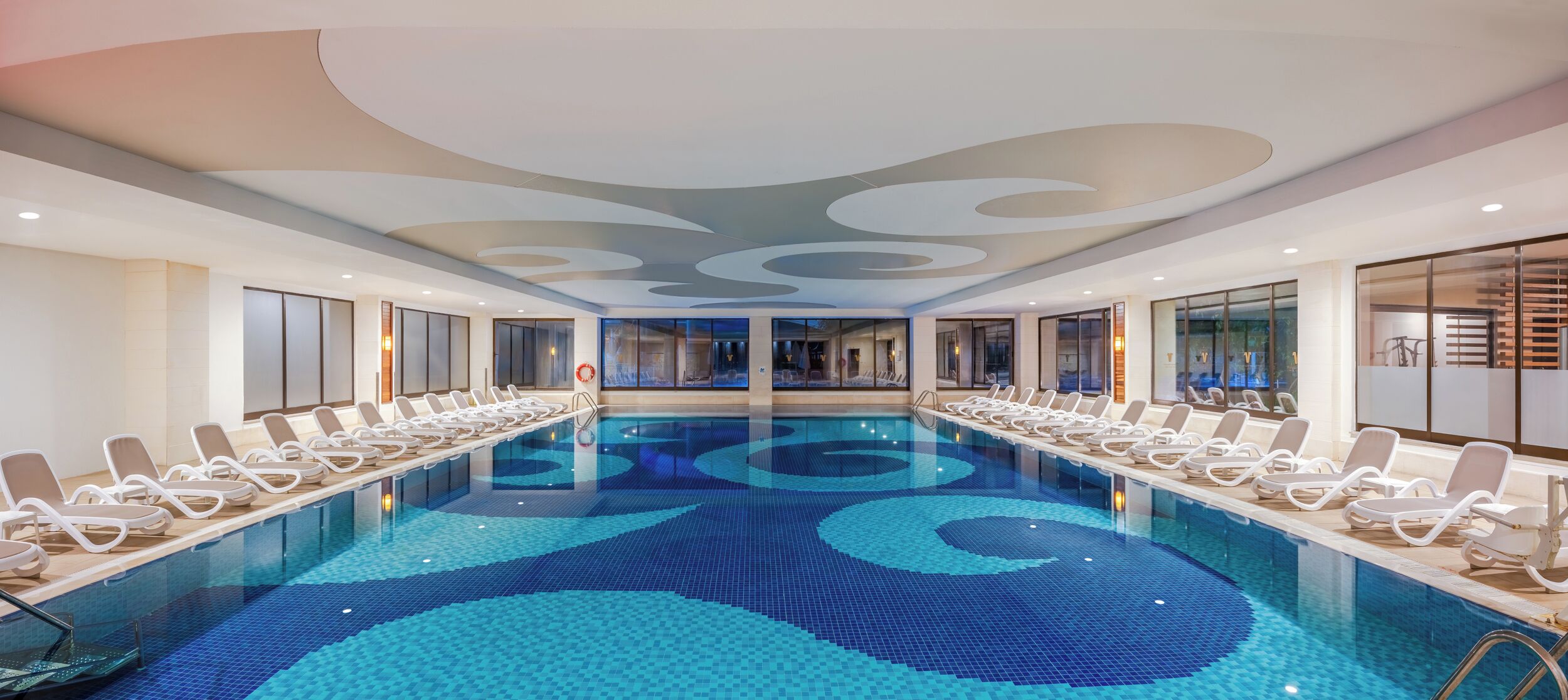 A large indoor swimming pool with lounge chairs along the sides, a modern ceiling design, and large windows.