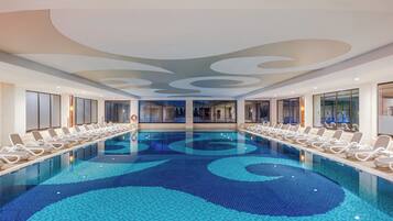 A large indoor swimming pool with lounge chairs along the sides, a modern ceiling design, and large windows.