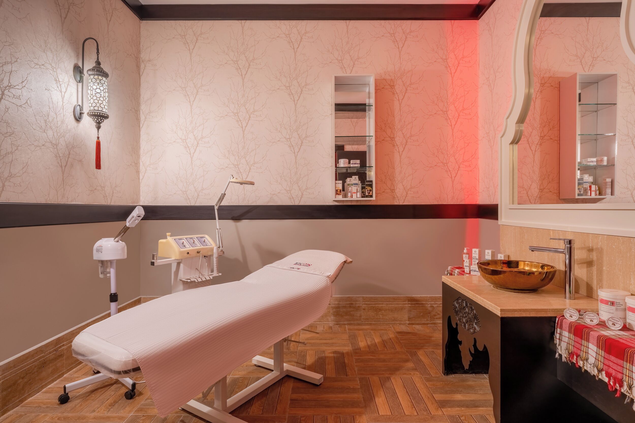 A medical examination room with a treatment bed, a wall-mounted lamp, a wooden floor, and a sink with a bowl.