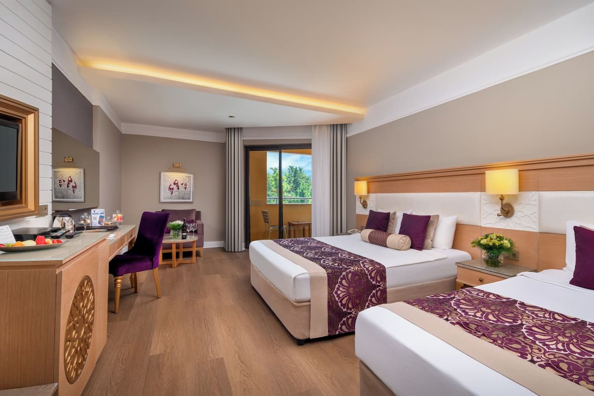A hotel room with two beds, a desk with a flat-screen TV, a chair, and a view of greenery through the window.